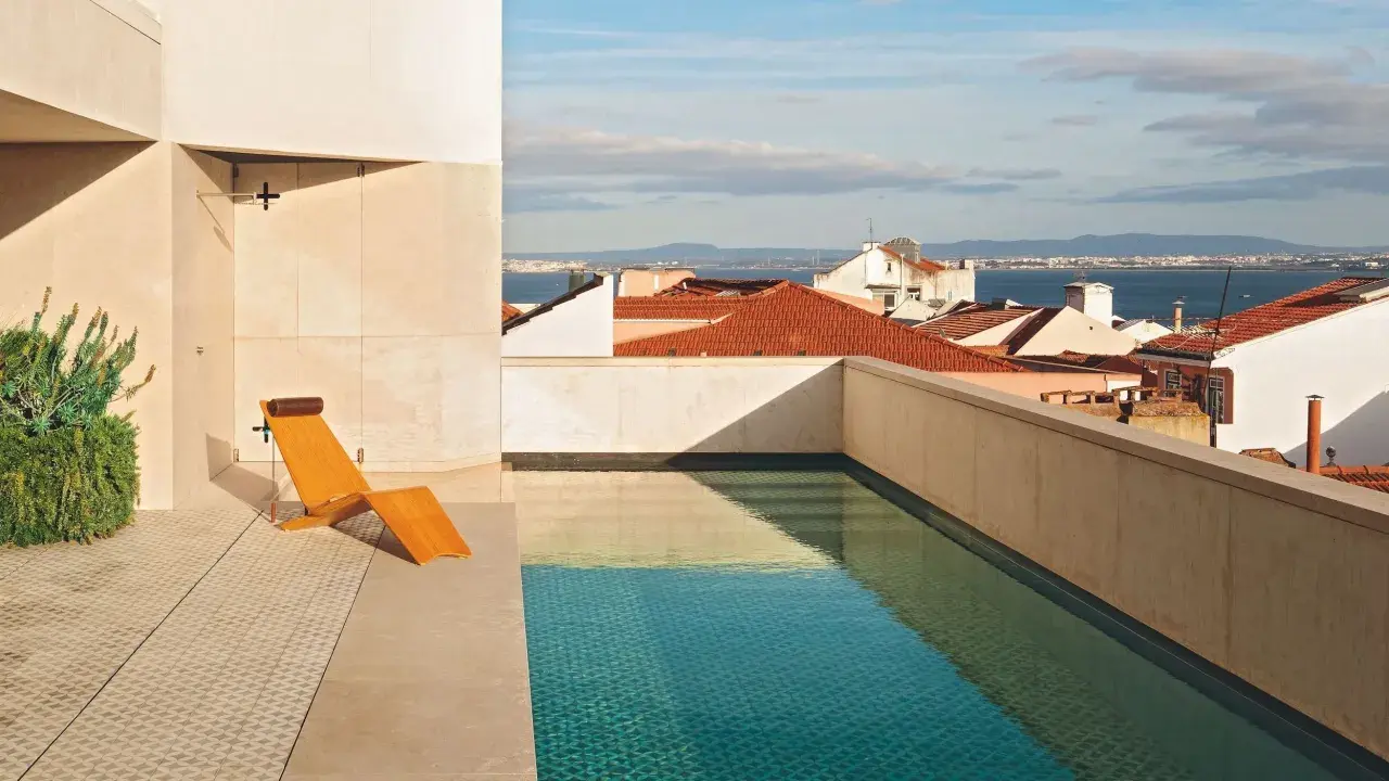 Una piscina en un balcón con vistas a tejados y al mar. Una tumbona de madera espera junto al agua cristalina.