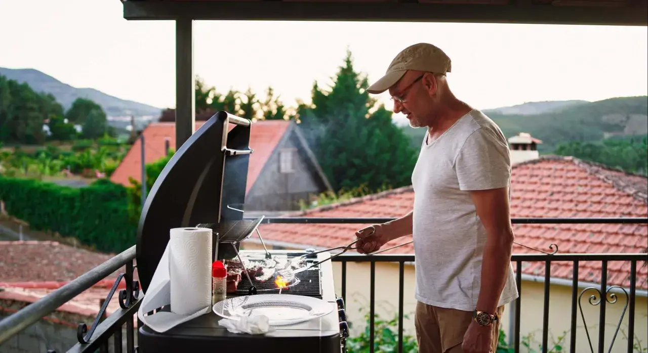 Hombre preparando barbacoa en patio interior con vistas a un pueblo.