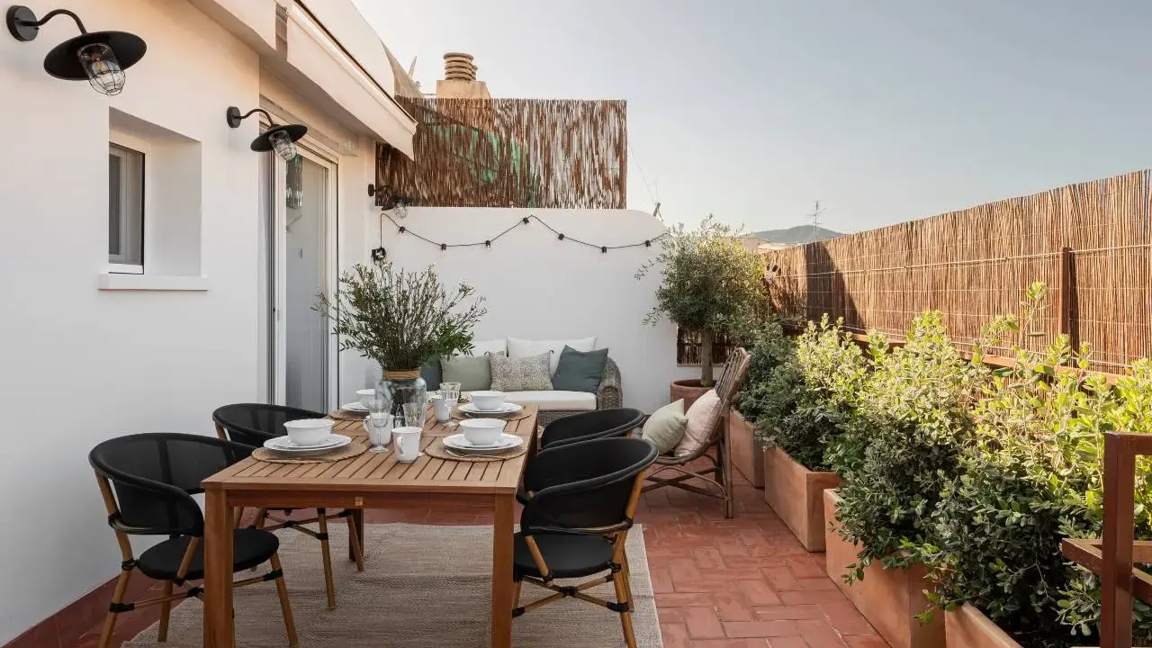 Acogedora terraza de 25 metros cuadrados con mesa de madera, sillas negras, sofá, plantas y luces decorativas.