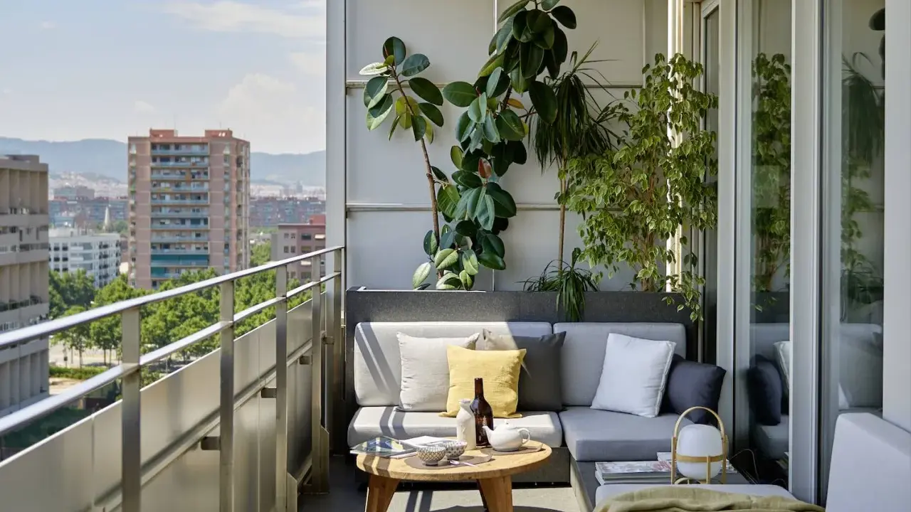 Acogedora terraza de una casa con sofá gris, plantas exuberantes y vistas urbanas. Ideal para relajarse al sol.