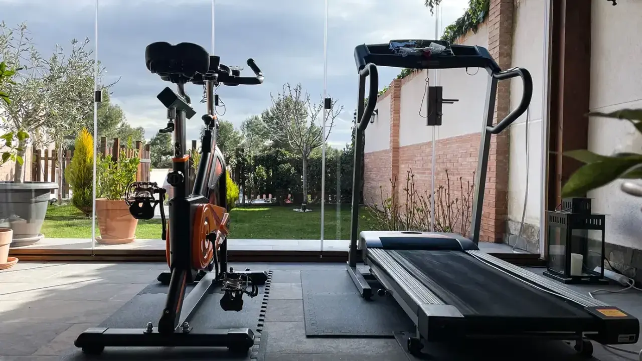 Un gimnasio en terraza con bicicleta estática y cinta de correr, con vistas a un jardín verde y un cielo nublado.