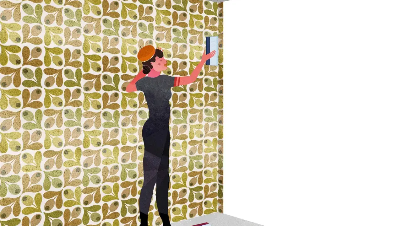 Mujer con boina naranja aplica papel tapiz con diseño de olivas. Muestra por dónde empezar a empapelar una pared, con herramientas en el suelo.
