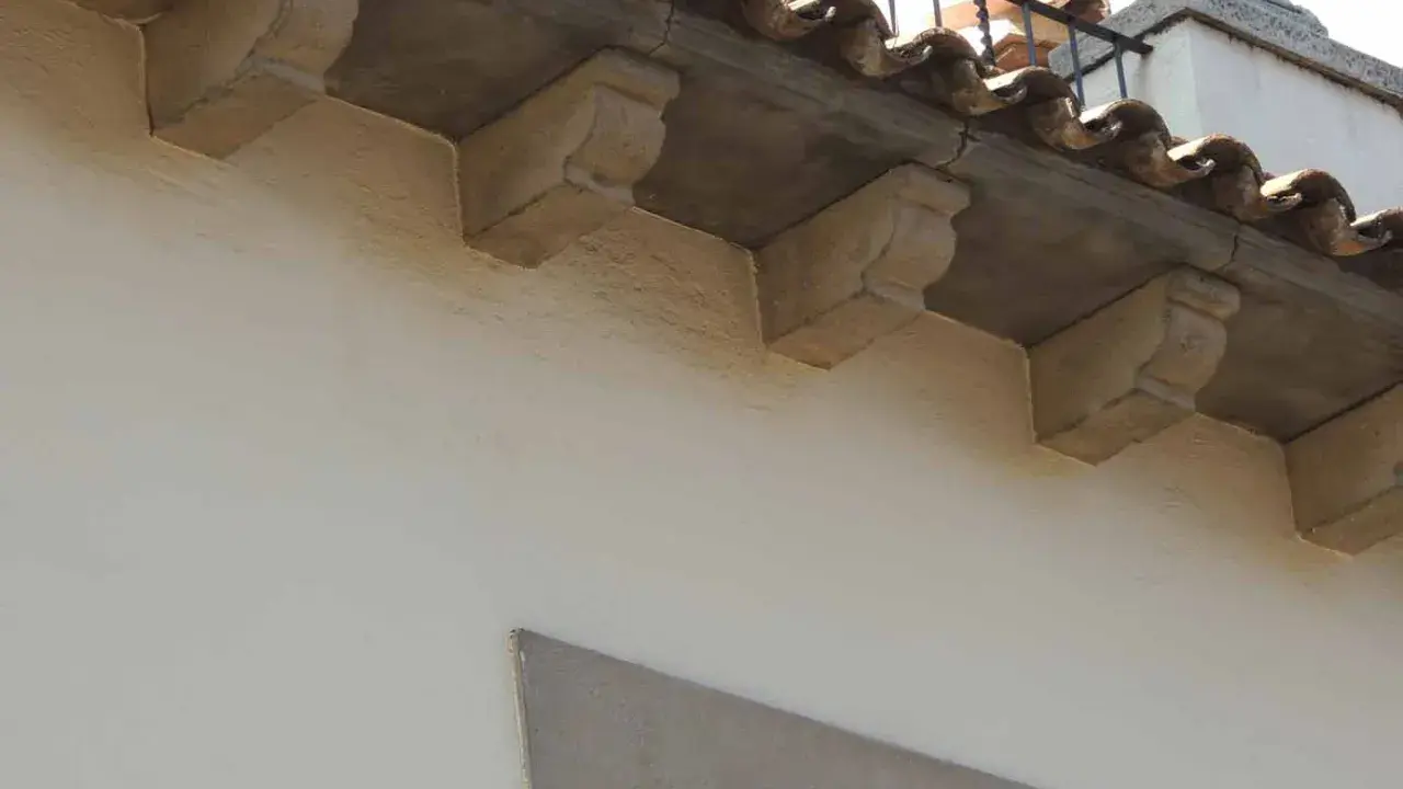 Detalle de qué es una cornisa, con ménsulas de piedra decorativas bajo tejas curvas y una barandilla metálica.