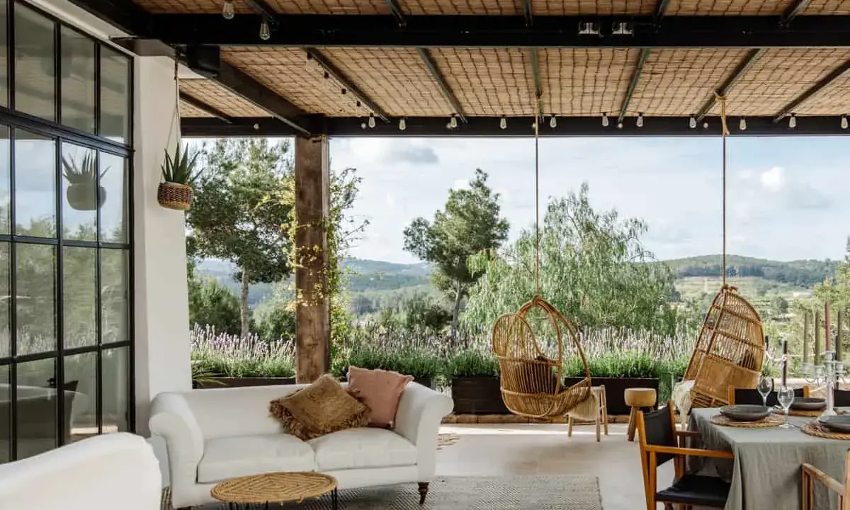 Terraza minimalista con sofá blanco, sillas colgantes de mimbre y vistas a un paisaje verde.