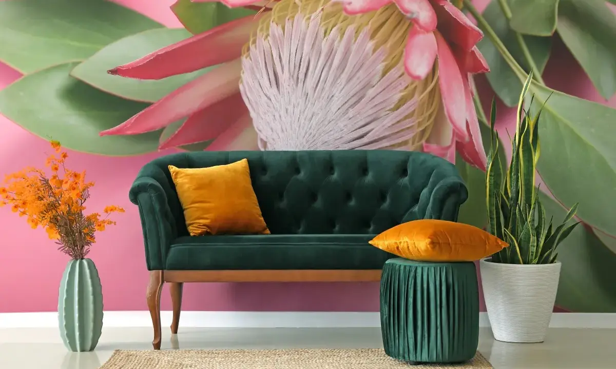 Sofá verde esmeralda con cojines naranjas, junto a un jarrón de cactus y plantas. El fondo es un mural con grandes estampados florales y hojas verdes sobre rosa vibrante.