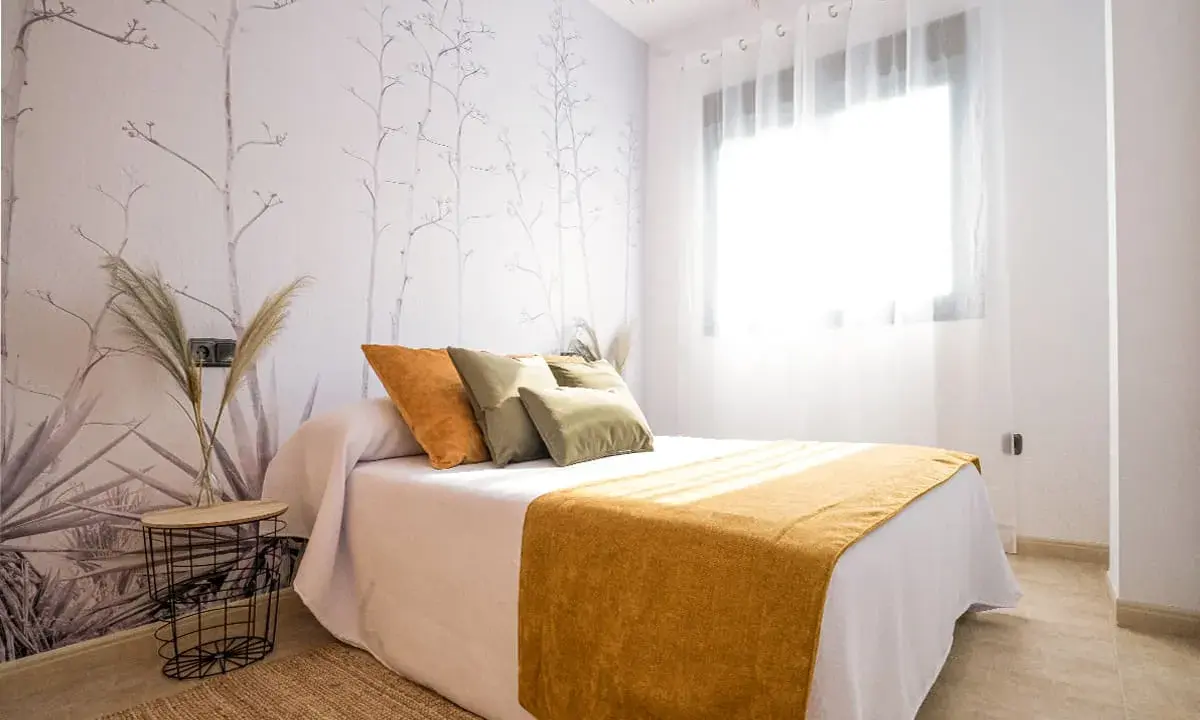 Habitación con papel tapiz botánico y cama con cojines naranjas y verdes. Una manta mostaza ilumina el espacio, ideal para quienes buscan colores para paredes con gotele.