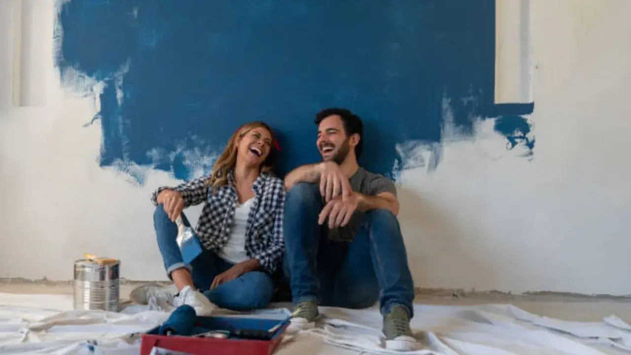 Pareja riendo sentada en el suelo frente a una pared recién pintada de azul. ¡Qué divertido es pintar una habitación!