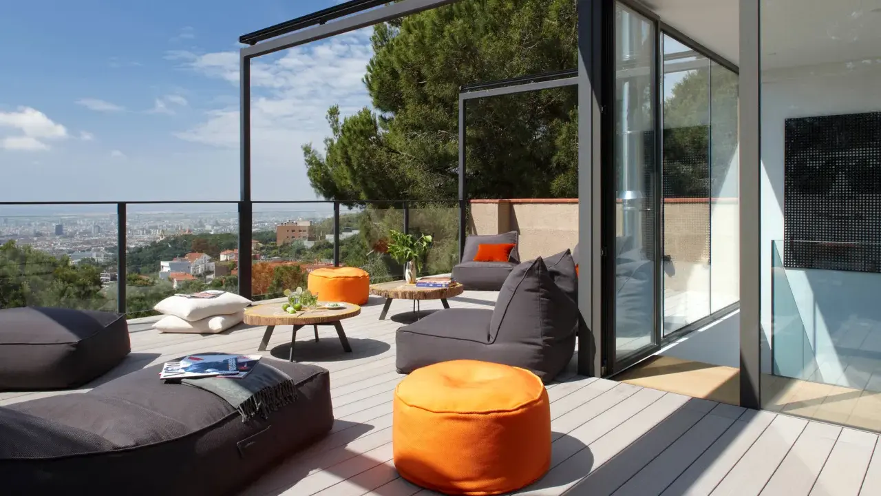 Cómodo balcón terraza con sillones, pufs naranjas y mesas bajas, con vistas a la ciudad.