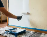 Hombre pintando una pared lisa de blanco con rodillo. Bandeja de pintura y brocha a la vista.