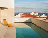 Una piscina en un balcón con vistas a tejados y al mar. Una tumbona de madera espera junto al agua cristalina.