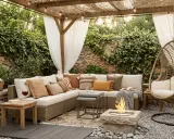 Acogedora zona chill out exterior con sofá modular, sillón colgante, hoguera y cortinas blancas. Un rincón perfecto para relajarse.