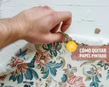 Mano retirando papel pintado floral de una pared blanca. Aprende cómo quitar papel pintado sin dañar la pared.