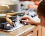 Mujer ajustando la perilla de la estufa de gas, con una sartén de verduras cocinándose. La llama azul indica que usa butano o gas natural.