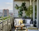 Acogedora terraza de una casa con sofá gris, plantas exuberantes y vistas urbanas. Ideal para relajarse al sol.