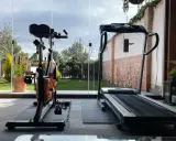 Un gimnasio en terraza con bicicleta estática y cinta de correr, con vistas a un jardín verde y un cielo nublado.