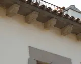 Detalle de qué es una cornisa, con ménsulas de piedra decorativas bajo tejas curvas y una barandilla metálica.