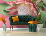 Sofá verde esmeralda con cojines naranjas, junto a un jarrón de cactus y plantas. El fondo es un mural con grandes estampados florales y hojas verdes sobre rosa vibrante.