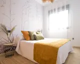 Habitación con papel tapiz botánico y cama con cojines naranjas y verdes. Una manta mostaza ilumina el espacio, ideal para quienes buscan colores para paredes con gotele.