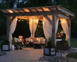Pérgola iluminada con luces cálidas y cortinas blancas, creando un ambiente acogedor. Una idea perfecta para saber como iluminar un jardin y disfrutarlo de noche.