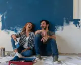 Pareja riendo sentada en el suelo frente a una pared recién pintada de azul. ¡Qué divertido es pintar una habitación!