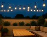 Ideas de iluminación para terraza: guirnaldas de bombillas cálidas iluminan un rincón acogedor con sofá y mesa de madera, creando un ambiente perfecto para la noche.