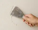 Mano con espátula rasca gotele de una pared, eliminando la textura irregular para prepararla.
