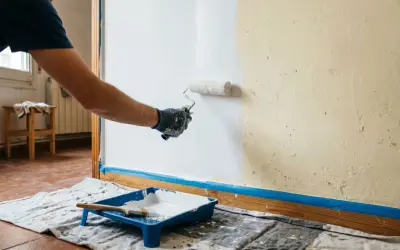 Hombre pintando una pared lisa de blanco con rodillo. Bandeja de pintura y brocha a la vista.