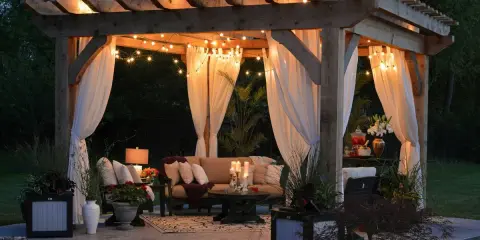 Pérgola iluminada con luces cálidas y cortinas blancas, creando un ambiente acogedor. Una idea perfecta para saber como iluminar un jardin y disfrutarlo de noche.