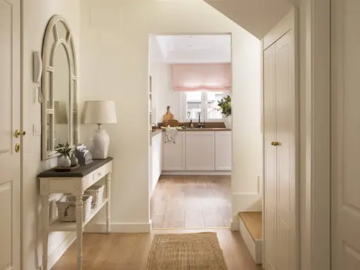 Un recibidor luminoso con espejo arqueado, consola con cestas y una cocina al fondo. ¡Ideal para pintar recibidor y darle un toque fresco!