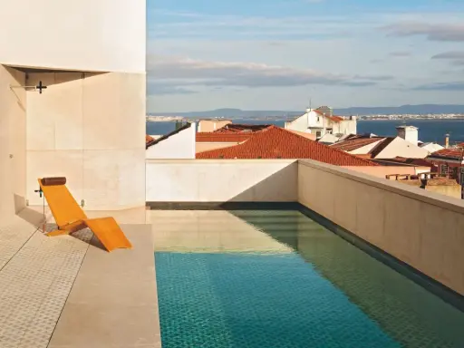 Una piscina en un balcón con vistas a tejados y al mar. Una tumbona de madera espera junto al agua cristalina.