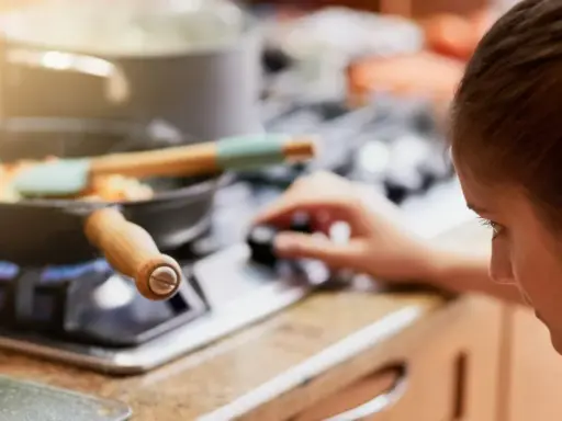 Mujer ajustando la perilla de la estufa de gas, con una sartén de verduras cocinándose. La llama azul indica que usa butano o gas natural.