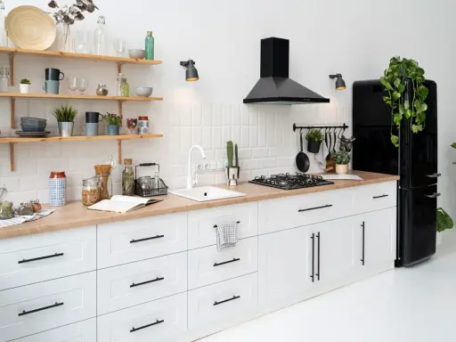 Cocina moderna con gabinetes blancos y encimera de madera. El diseño para pintar cocinas es minimalista, con electrodomésticos negros y estantes de madera.