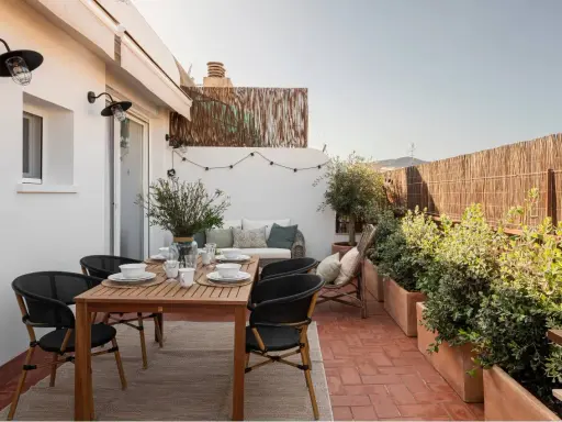 Acogedora terraza de 25 metros cuadrados con mesa de madera, sillas negras, sofá, plantas y luces decorativas.