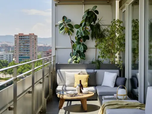 Acogedora terraza de una casa con sofá gris, plantas exuberantes y vistas urbanas. Ideal para relajarse al sol.