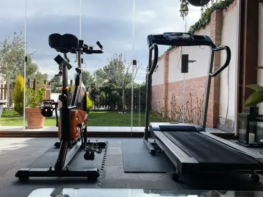 Un gimnasio en terraza con bicicleta estática y cinta de correr, con vistas a un jardín verde y un cielo nublado.
