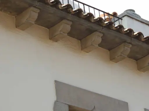 Detalle de qué es una cornisa, con ménsulas de piedra decorativas bajo tejas curvas y una barandilla metálica.