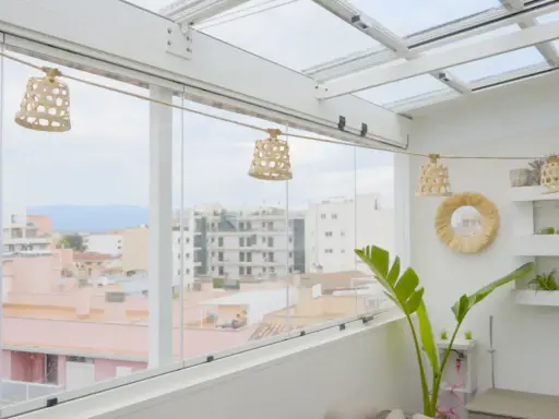 Un cerramiento ático con vistas urbanas, decorado con lámparas de mimbre y plantas.