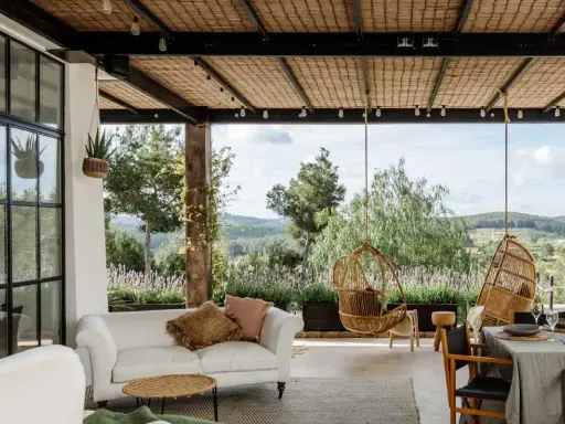 Terraza minimalista con sofá blanco, sillas colgantes de mimbre y vistas a un paisaje verde.