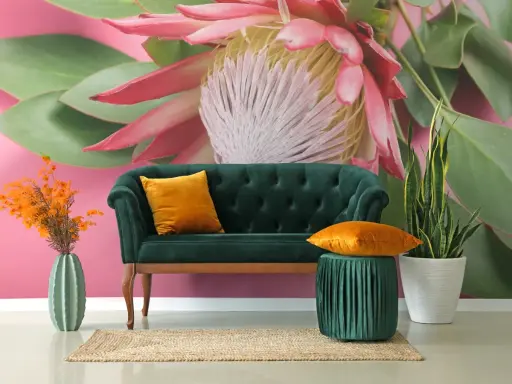 Sofá verde esmeralda con cojines naranjas, junto a un jarrón de cactus y plantas. El fondo es un mural con grandes estampados florales y hojas verdes sobre rosa vibrante.