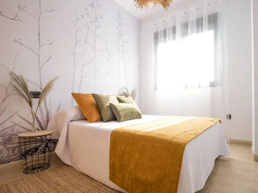 Habitación con papel tapiz botánico y cama con cojines naranjas y verdes. Una manta mostaza ilumina el espacio, ideal para quienes buscan colores para paredes con gotele.