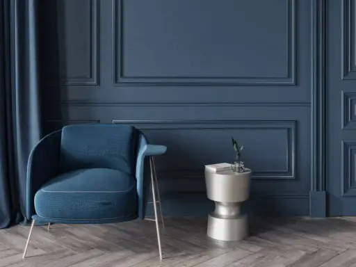 Elegante sillón azul junto a una mesa auxiliar plateada, con cortinas a juego y detalladas molduras en paredes de tono oscuro.