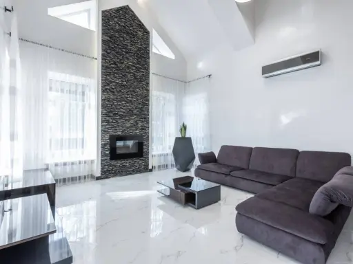 Un moderno salón con un sofá gris oscuro, chimenea de piedra y suelo de mármol blanco.