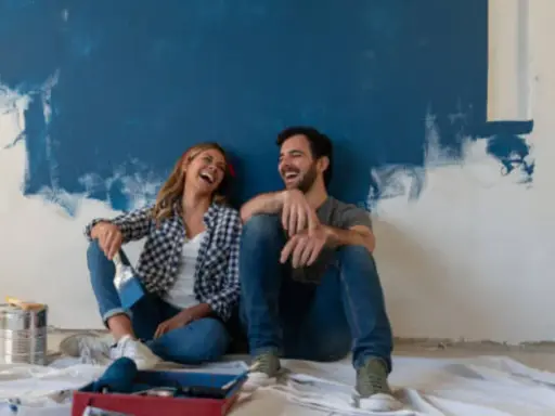 Pareja riendo sentada en el suelo frente a una pared recién pintada de azul. ¡Qué divertido es pintar una habitación!