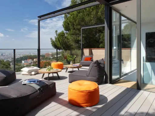 Cómodo balcón terraza con sillones, pufs naranjas y mesas bajas, con vistas a la ciudad.