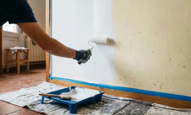 Hombre pintando una pared lisa de blanco con rodillo. Bandeja de pintura y brocha a la vista.