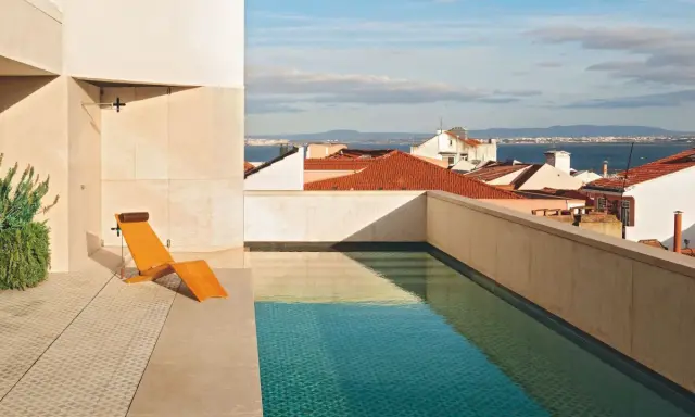 Una piscina en un balcón con vistas a tejados y al mar. Una tumbona de madera espera junto al agua cristalina.