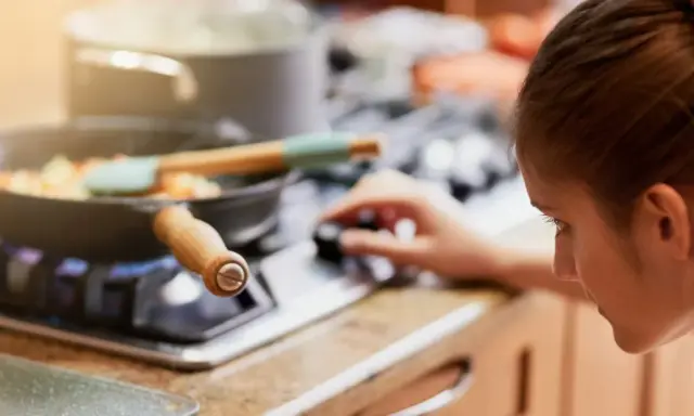 Mujer ajustando la perilla de la estufa de gas, con una sartén de verduras cocinándose. La llama azul indica que usa butano o gas natural.