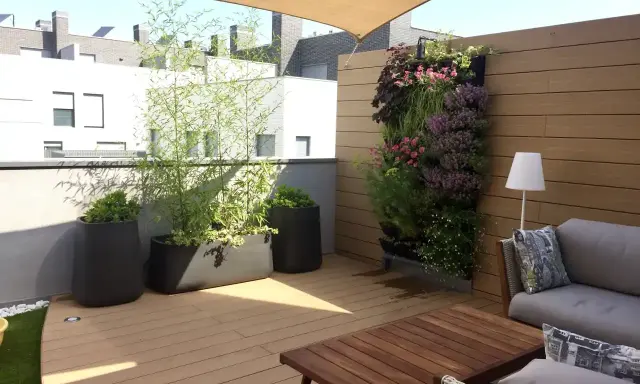 Un **jardín moderno** en una terraza con sofá, mesa de madera, plantas en macetas y un muro verde vibrante.