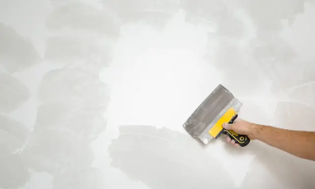 Mano con espátula trabajando en pared rugosa, como quitar pintura.