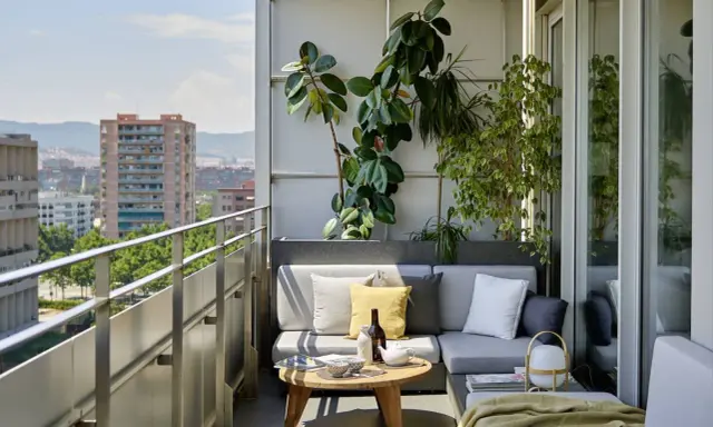 Acogedora terraza de una casa con sofá gris, plantas exuberantes y vistas urbanas. Ideal para relajarse al sol.